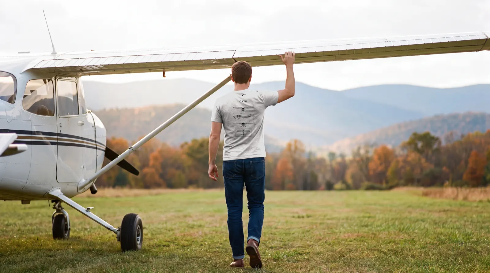 Pilot walking under the wing of a parked high wing trainer, one hand on the trailing edge, autumn ridges behind, back to the camera, wearing the C172 shirt in light grey