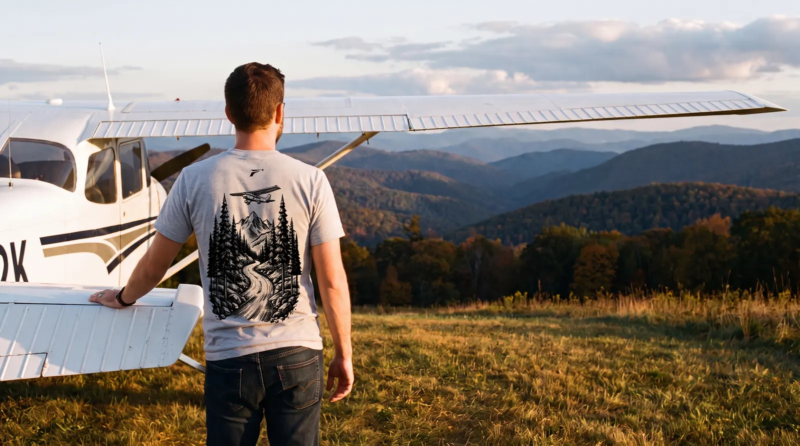 Pilot at the tail of a high wing trainer on a high grass field at golden hour looking out over layered ridges, back to the camera, the black Mountains print filling the back of the light grey shirt